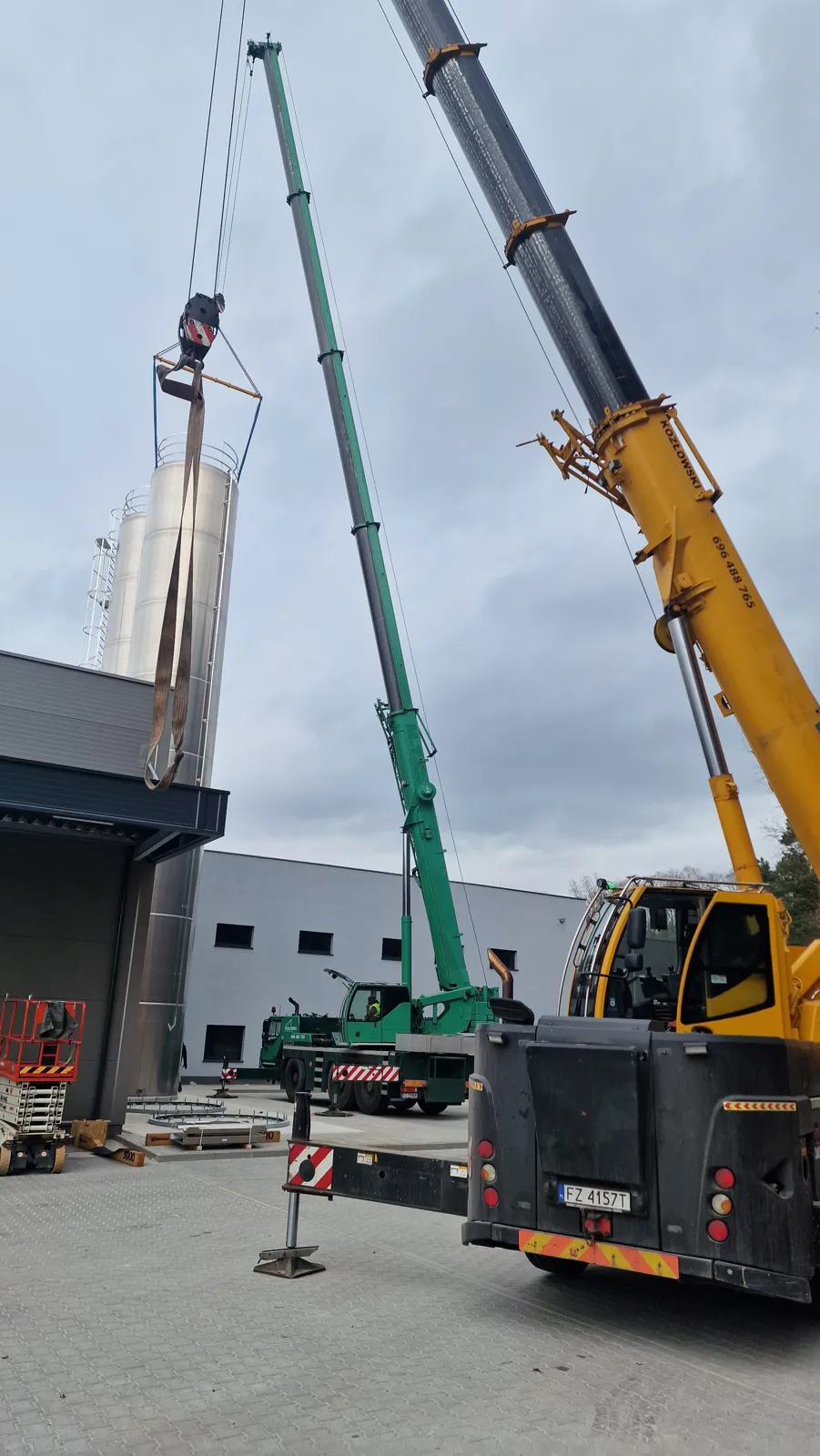 Two mobile cranes lifting a stainless steel silo in tandem